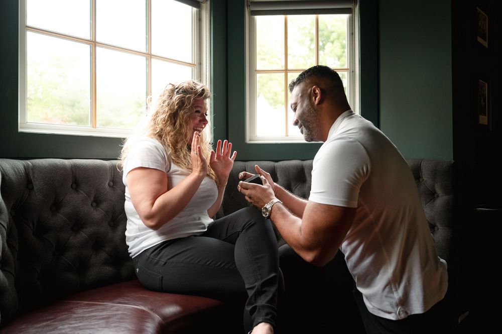 Two people sharing an emotional moment on a dark gray couch by a window during a proposal.