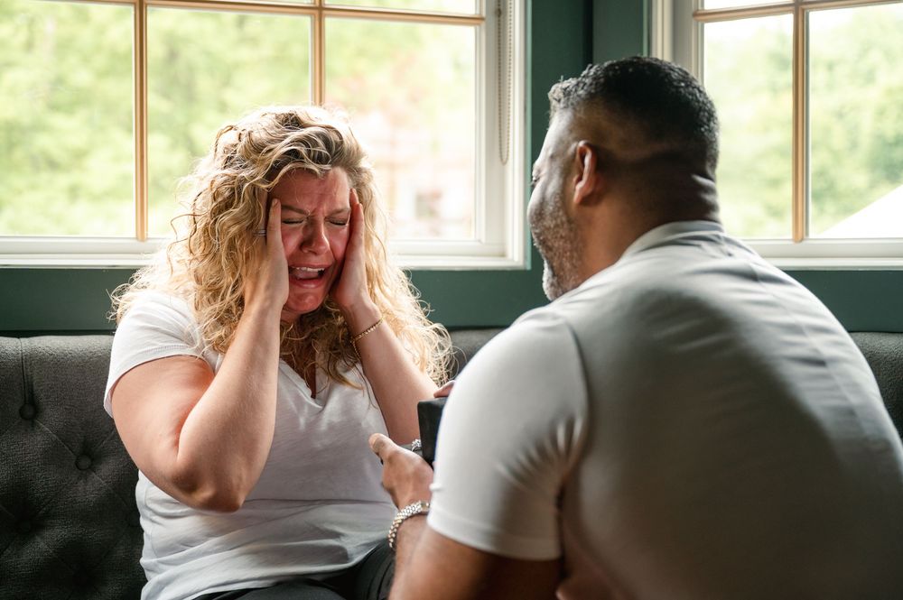 Two people sit on a couch in conversation with one appearing distressed while the other listens supportively.
