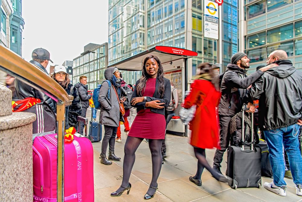Travelers with luggage wait at a busy urban street corner in cold weather with people bundled in coats.