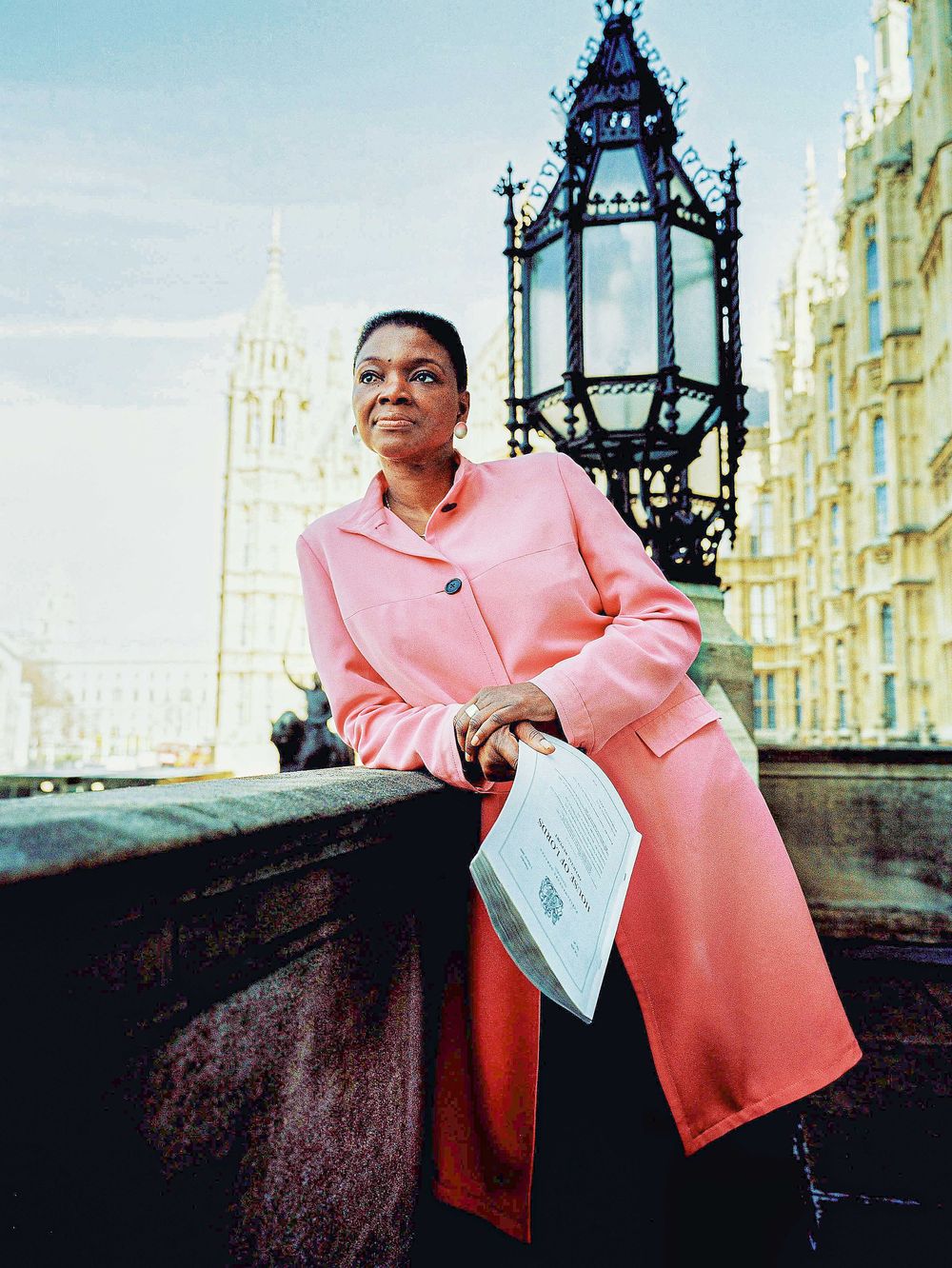 A person in a stylish pink coat stands near an ornate Victorian lamp post against historic London architecture.
