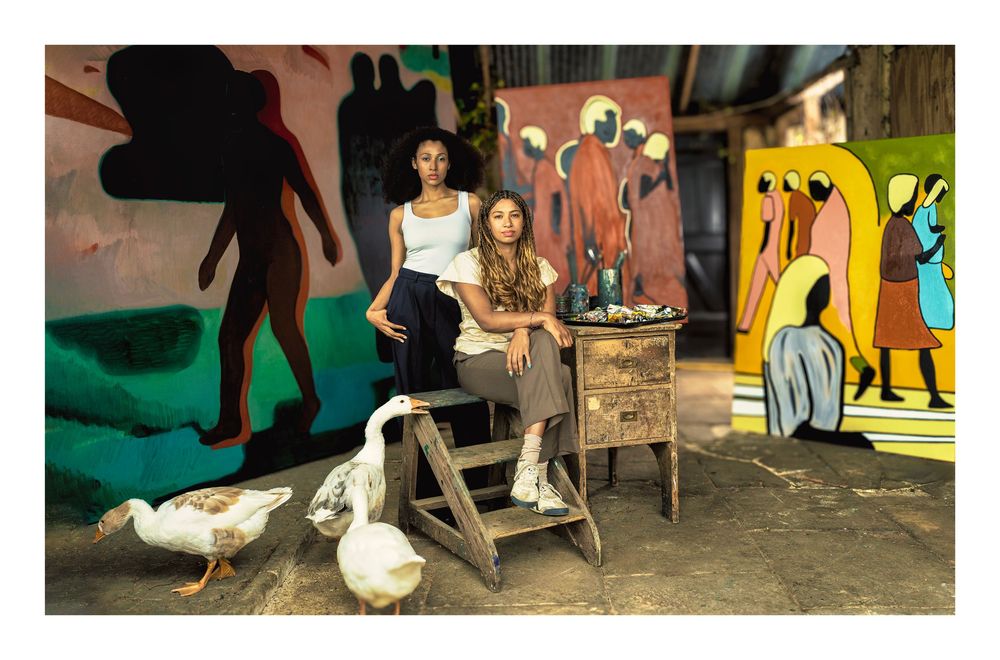 Woman seated on wooden chair in urban art space with white geese walking around her and colorful murals on walls.