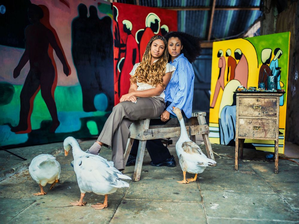 Two people sit on a bench near colorful graffiti walls while geese waddle around their feet in an urban setting.