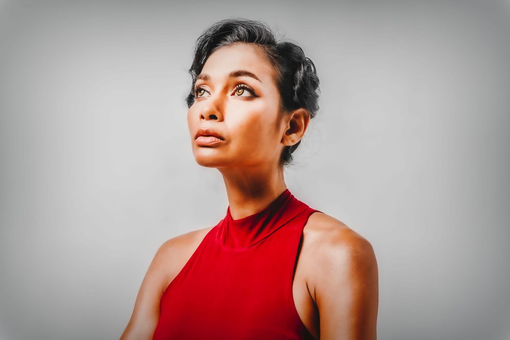 Person in a sleeveless red top poses against gray background with dramatic lighting and upward gaze.