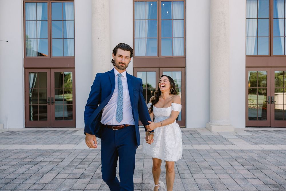 Two people in formal attire laugh together against a backdrop of white pillars.