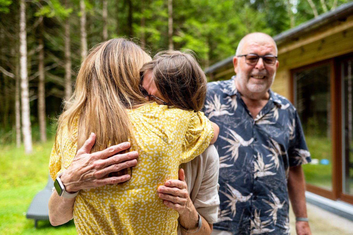 Two people share an embrace outdoors while another person in a floral shirt smiles in the background.