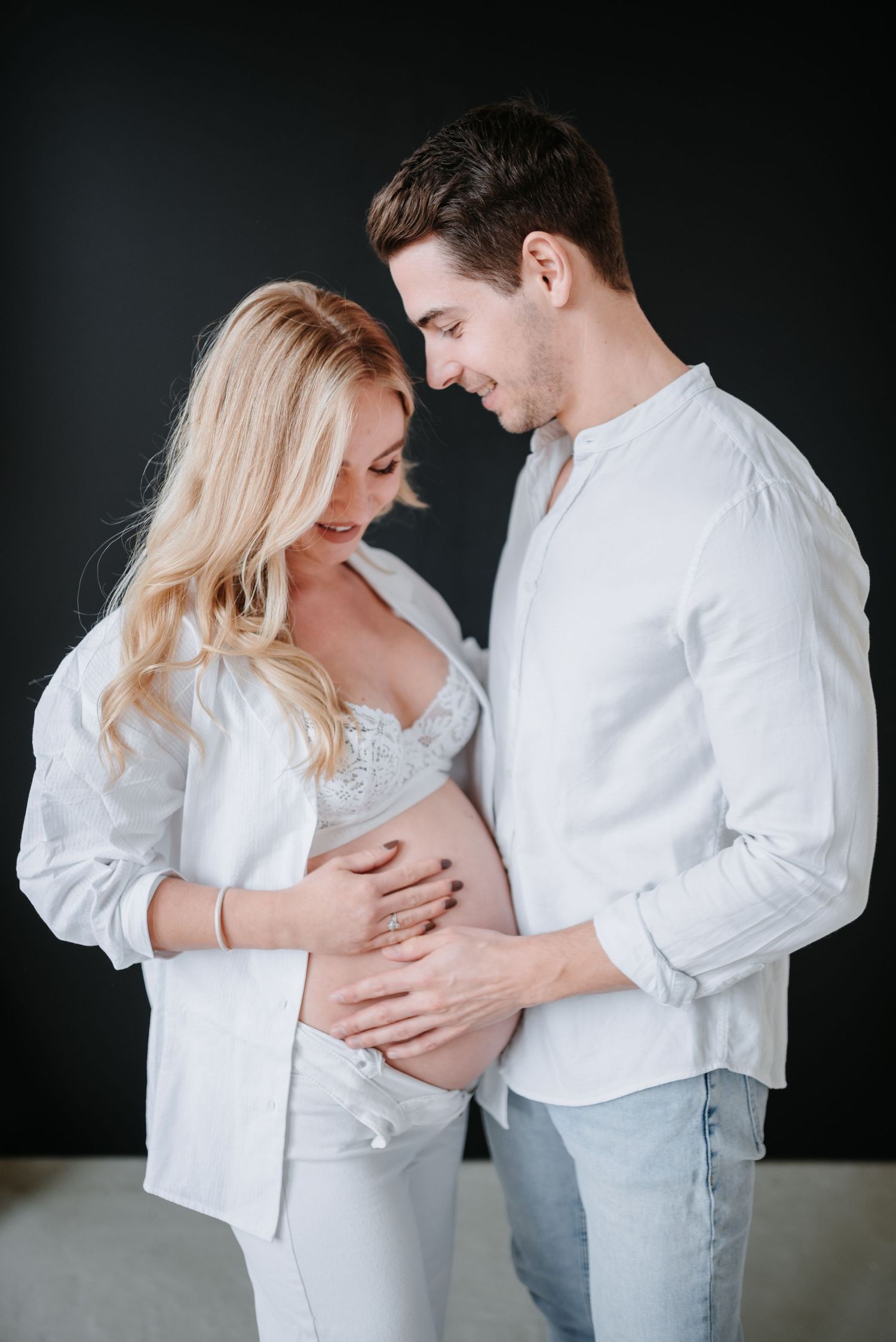 Intimate maternity portrait of couple in white shirts embracing against dark background during pregnancy photoshoot.