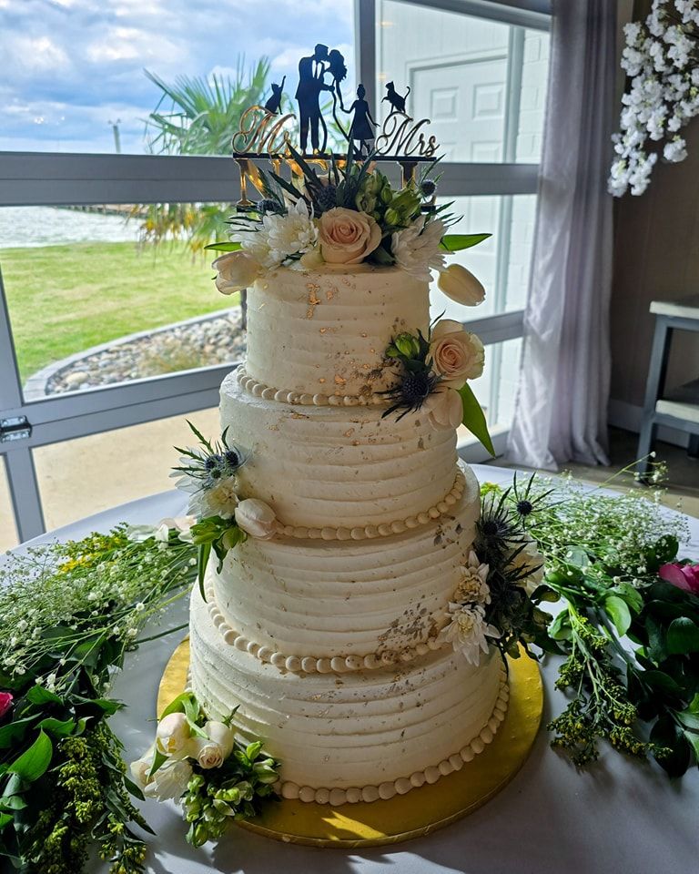 A four-tier white buttercream wedding cake decorated with roses and greenery sits by a window overlooking the ocean.