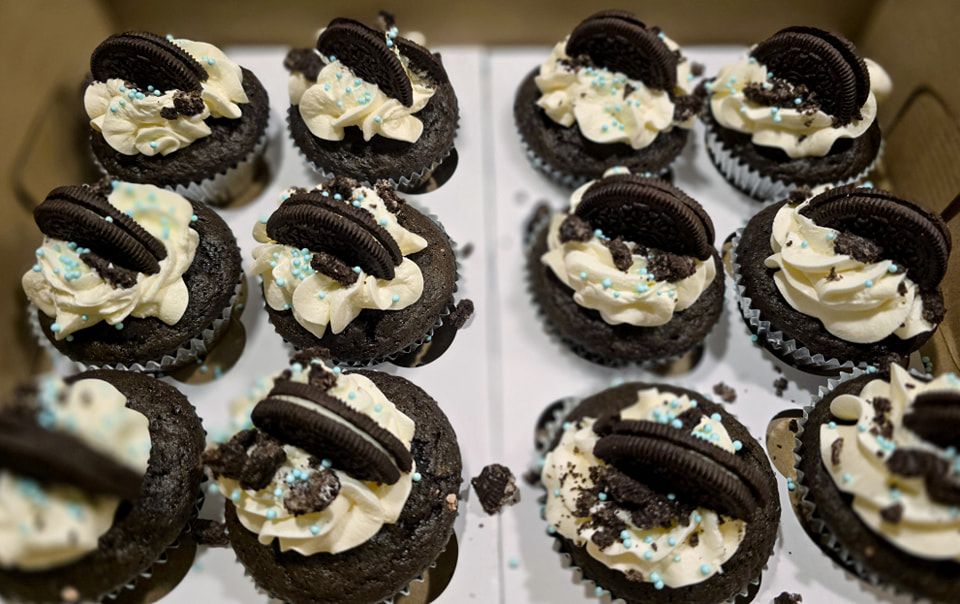 A dozen chocolate Oreo cupcakes with white frosting swirls and cookie pieces on top.