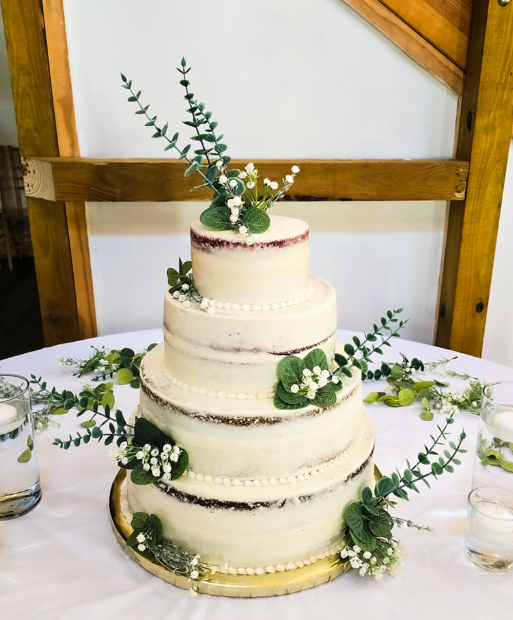 Four-tier white semi-naked wedding cake decorated with fresh greenery and small white flowers on a golden cake board.