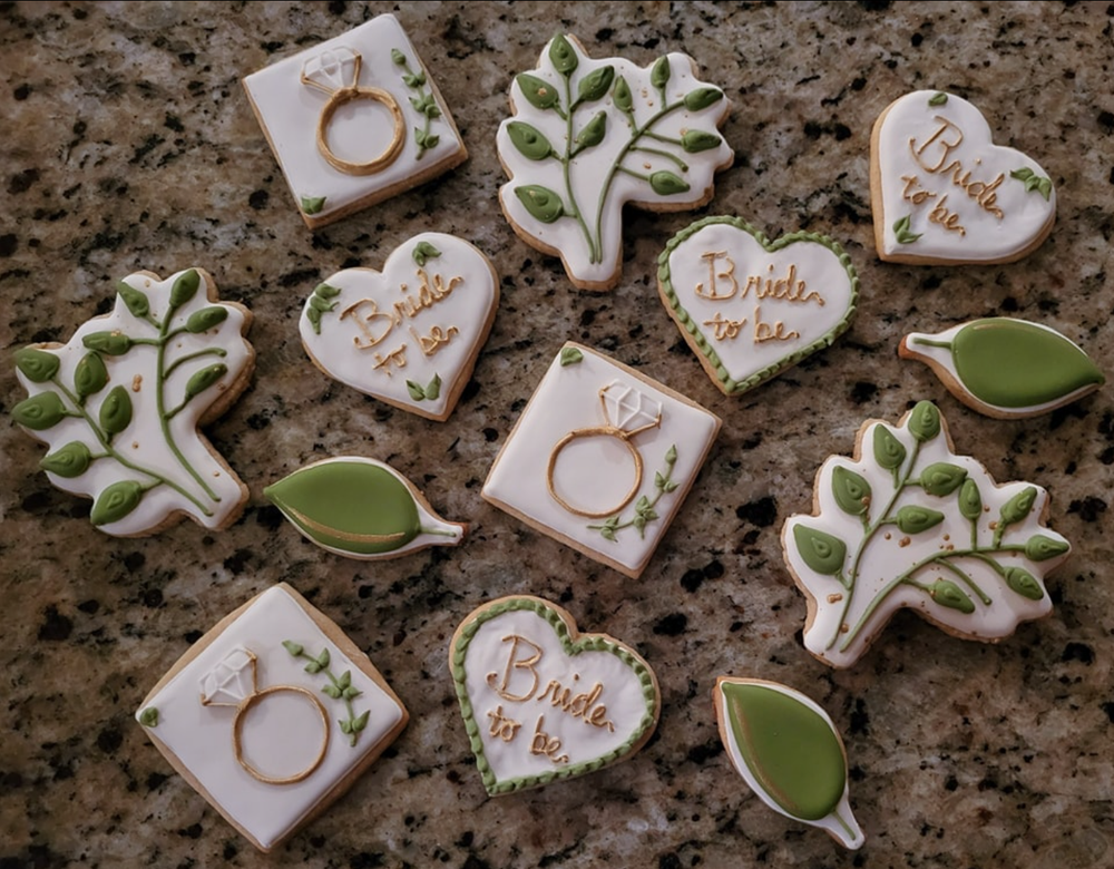 Decorated sugar cookies with bridal designs including rings, hearts, and leaves arranged on a dark granite countertop.