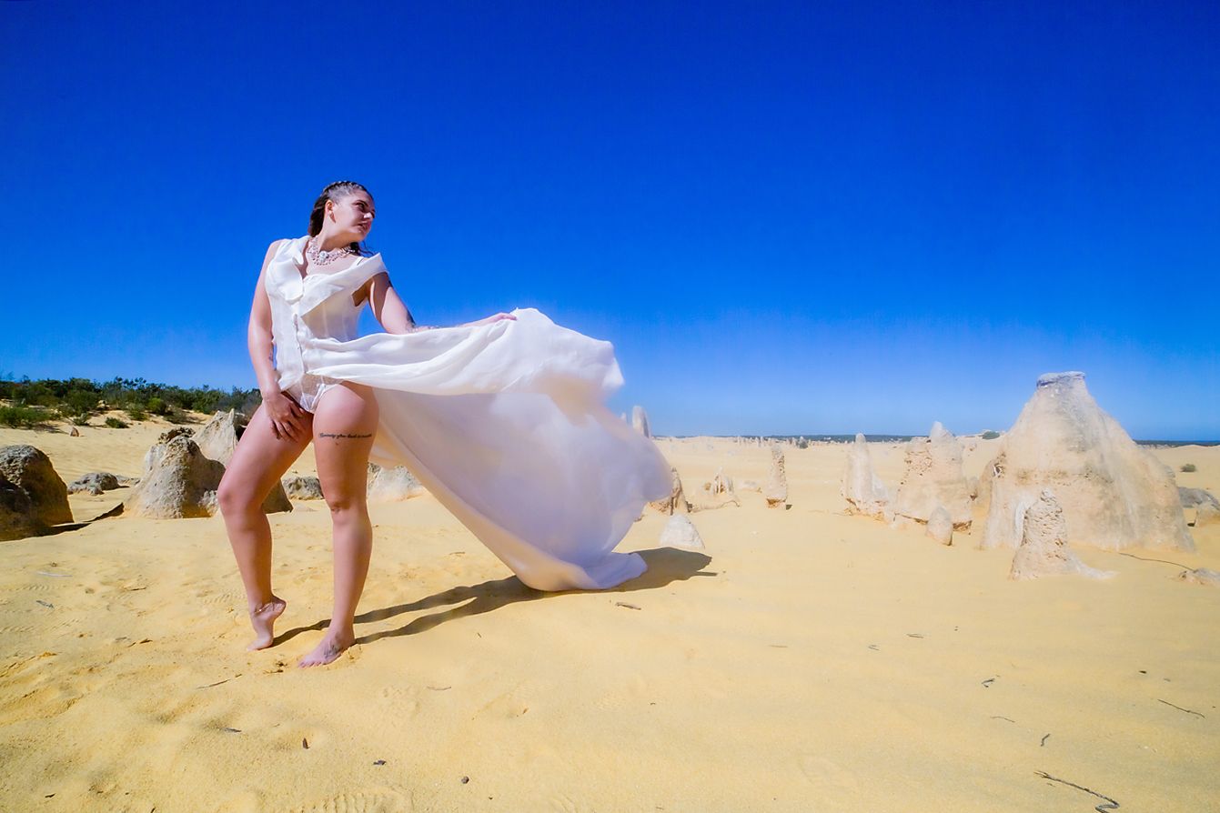 A flowing white dress billows in the wind against a sandy landscape and bright blue sky at Pinnacles Desert.
