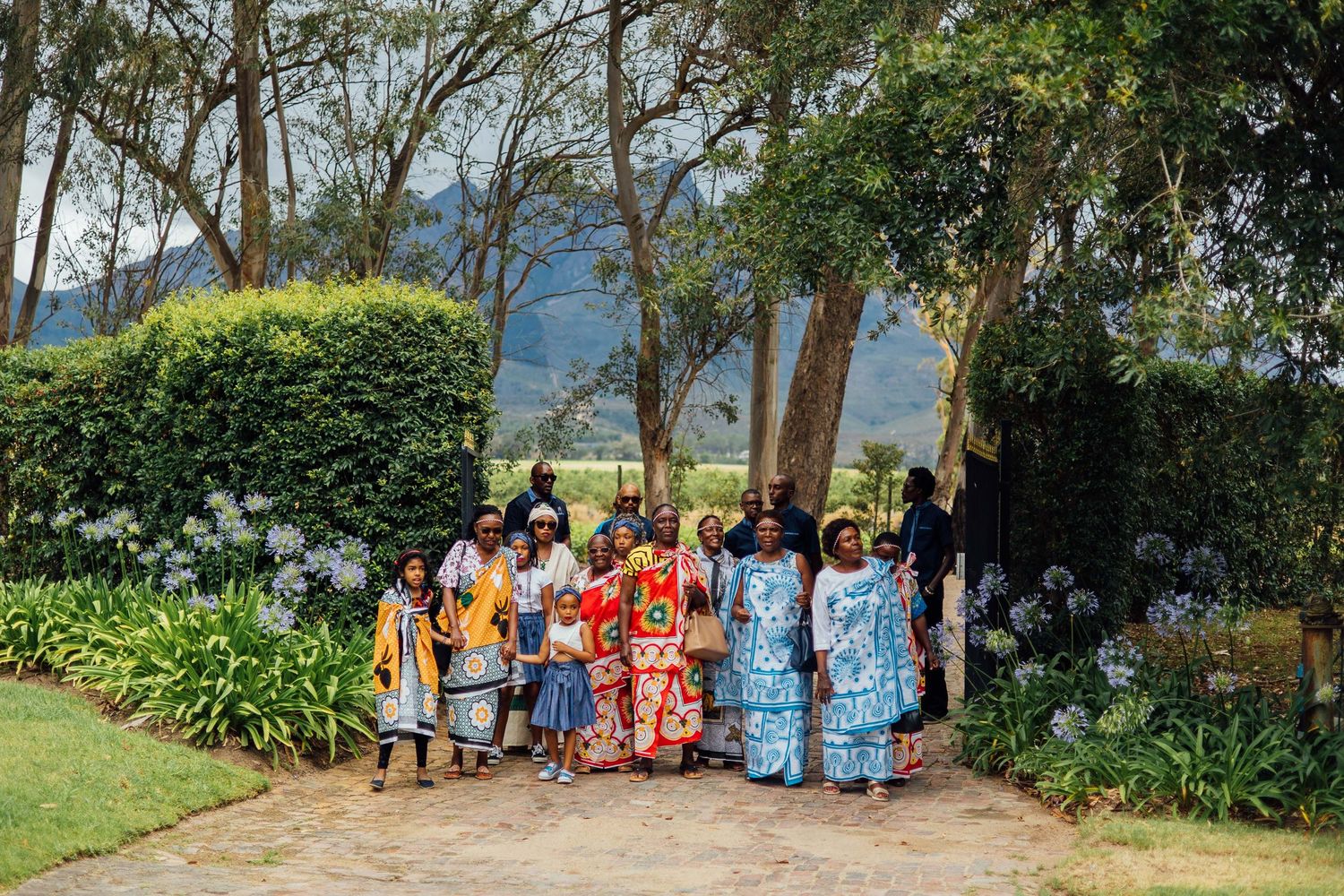 A diverse group wearing traditional African clothing gathers outdoors for a group photo near trees and greenery.