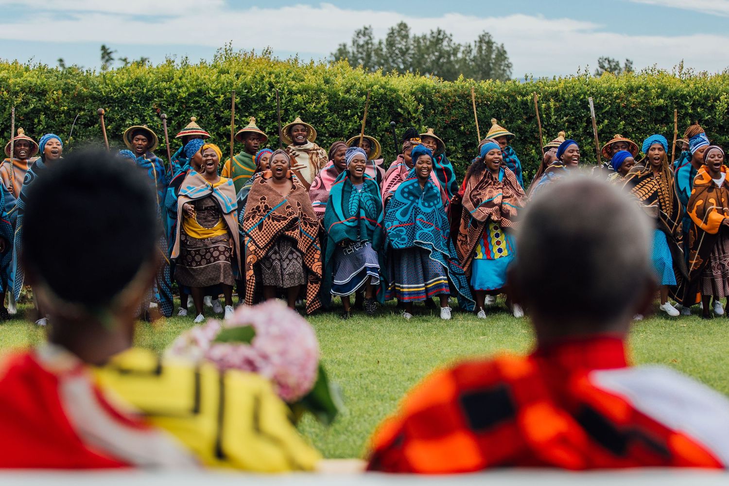 People wearing colorful traditional clothing gather outdoors in front of green hedges during a cultural celebration.