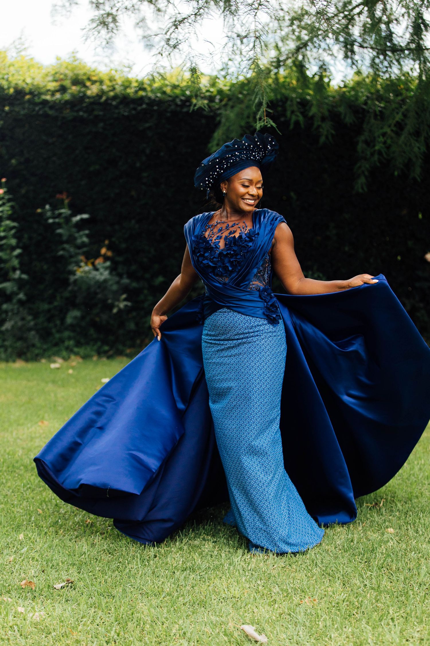 A flowing royal blue formal gown with a fitted bodice and dramatic cape-like overlay twirls on a green lawn.