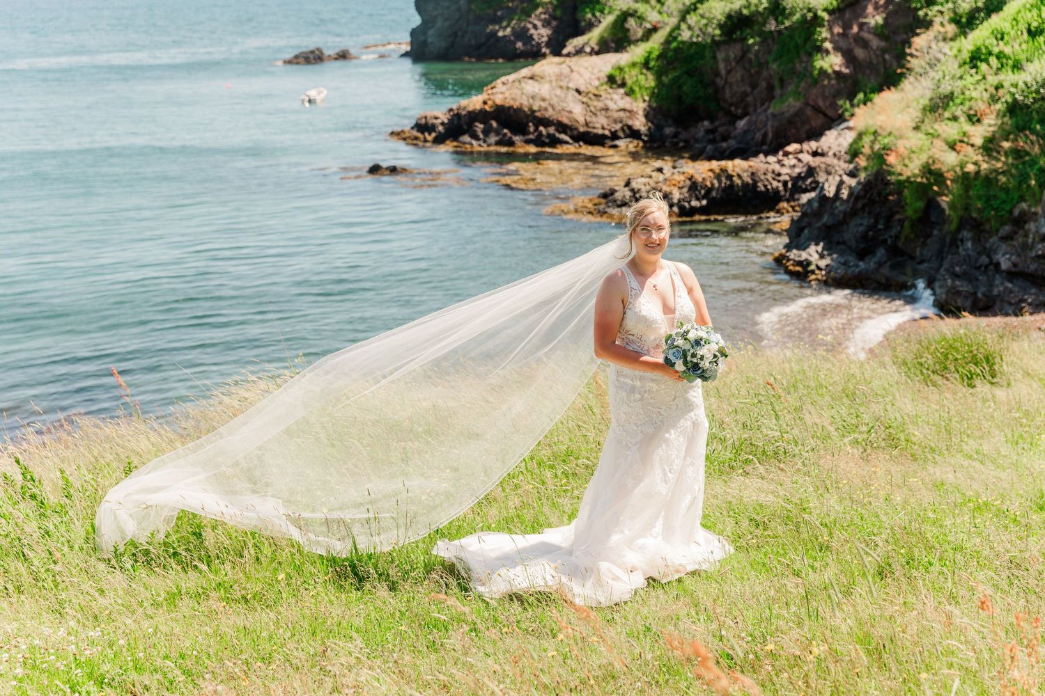 A flowing wedding dress catches the breeze along a scenic coastal cliff overlooking blue ocean waters.