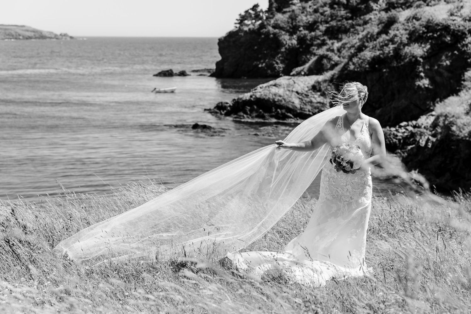 A bride in a flowing white gown poses dramatically against a scenic coastal cliffside landscape.