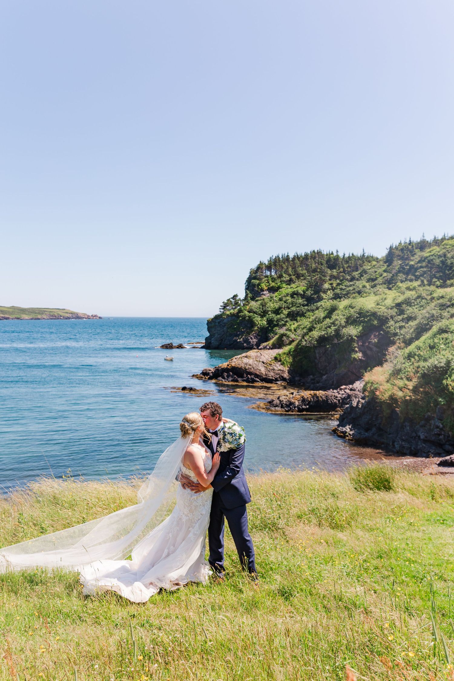 A couple shares an intimate moment on a grassy seaside cliff overlooking the blue ocean on a sunny day.