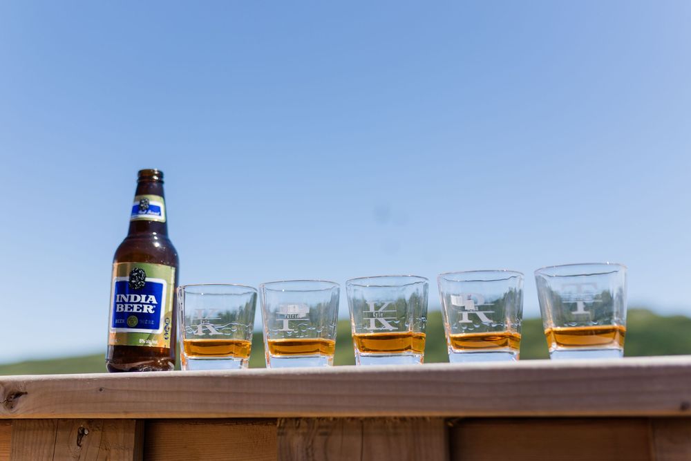 Line of whiskey shots on wooden deck railing with Samuel Adams beer bottle against blue sky background.