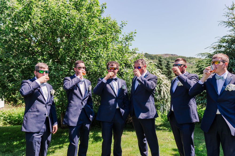A group in formal navy suits and dress shirts gather outdoors for a wedding photo shoot in a sunny garden setting.