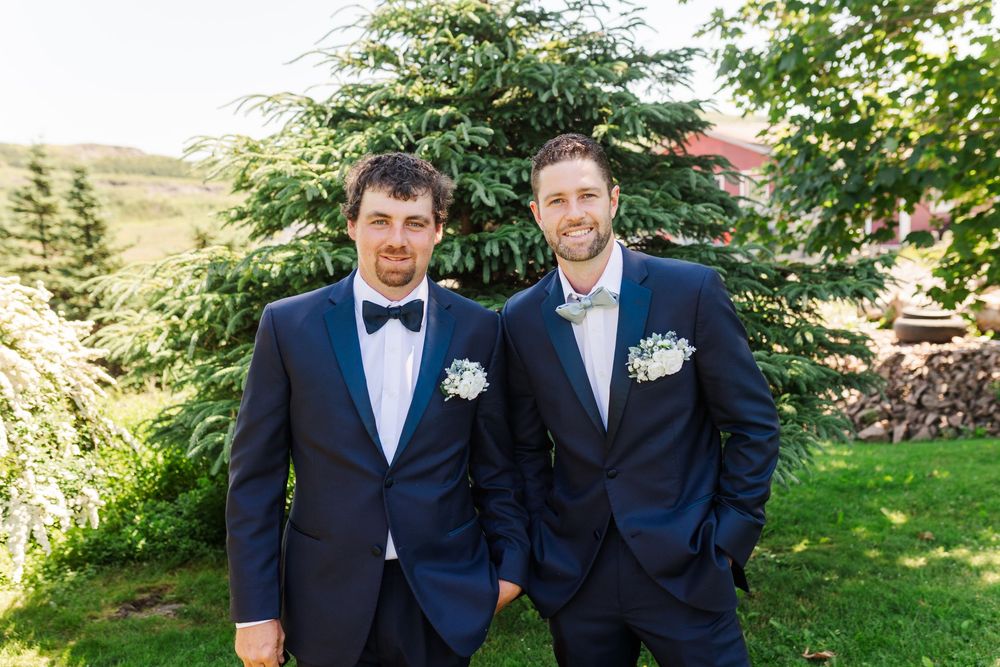Two people in navy blue suits and boutonnieres stand together outdoors in a garden setting on a sunny day.