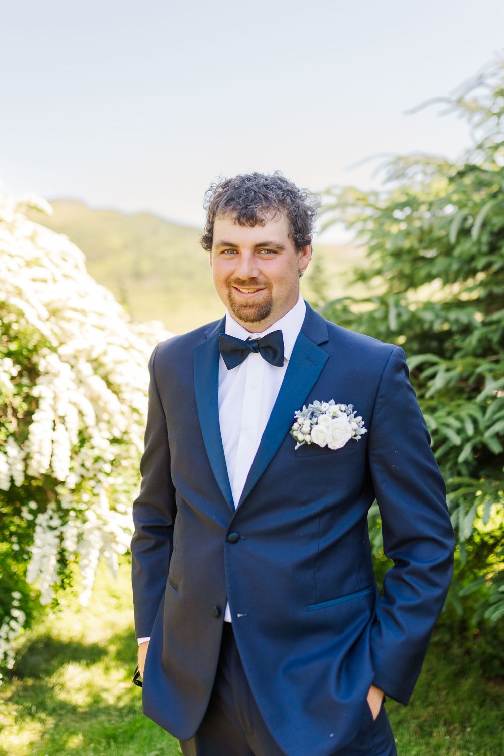 A wedding portrait outdoors with a navy blue suit and bowtie, featuring a white floral boutonniere and natural sunlit backdrop.