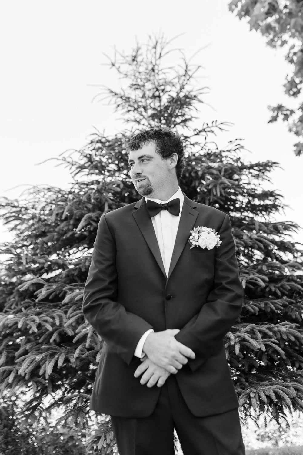 Wedding party members in black tuxedos stand together outdoors against an evergreen tree background.