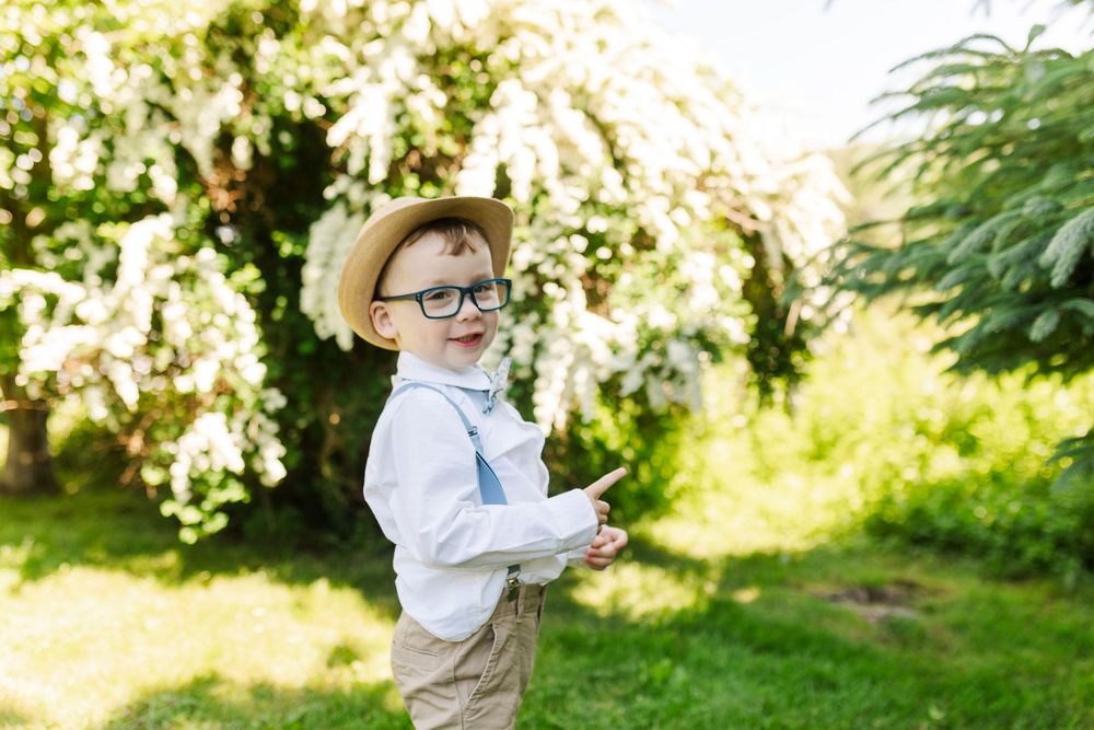 Young boy in button down shirt explores nature outdoors in spring garden with flowering trees and greenery.