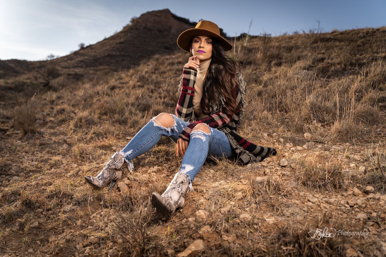 A rustic western style photo of someone sitting on a hillside wearing boots, jeans, and a wide-brimmed hat.