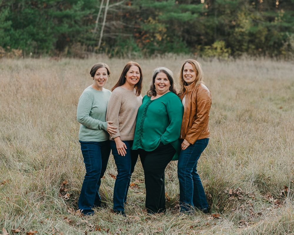 Four women in casual attire laughing and embracing while standing together in a grassy field.
