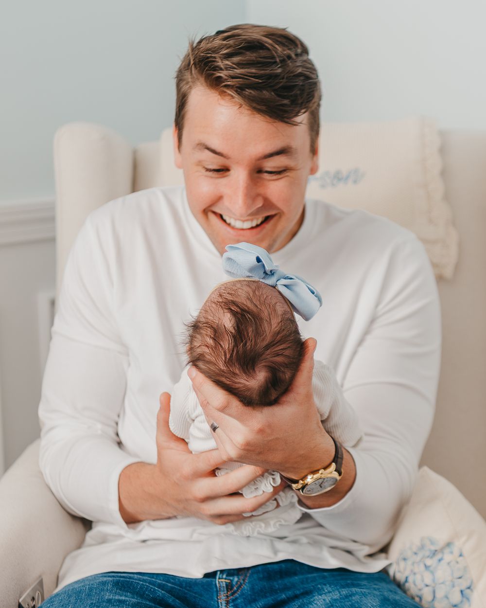 A tender parenting moment showing someone in a white sweater holding a tiny baby with a blue bow on beige chair.
