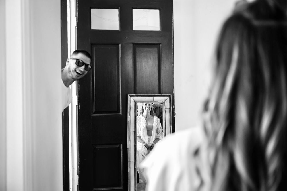 A black and white photo of someone playfully peeking around a doorway while another person looks in a mirror.