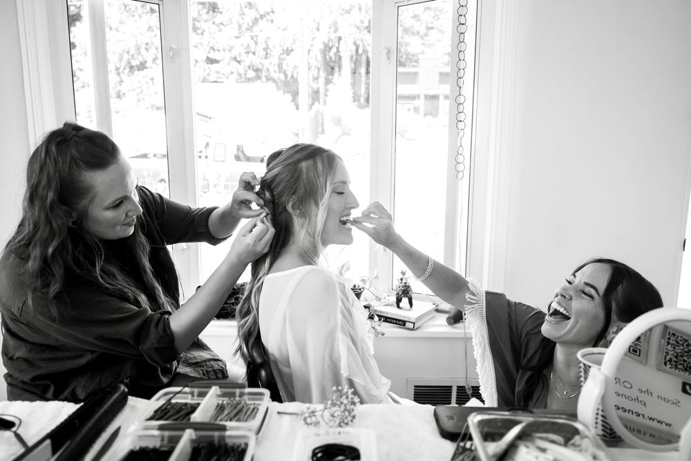 Black and white candid image of people getting ready with hair and makeup near a bright window with laughter and joy.