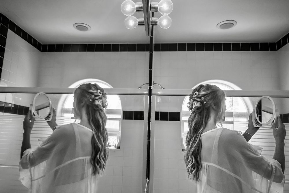 Black and white reflection in bathroom mirror shows long-haired figure in white robe reaching up to adjust their hair.