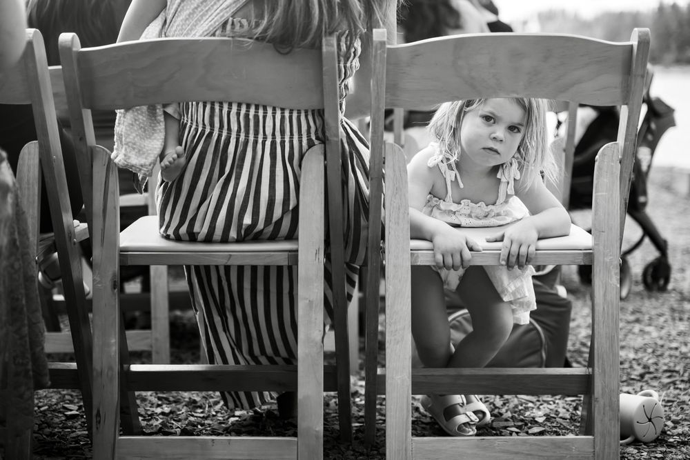Black and white photograph showing child peeking through a gap between wooden folding chairs at an outdoor event.