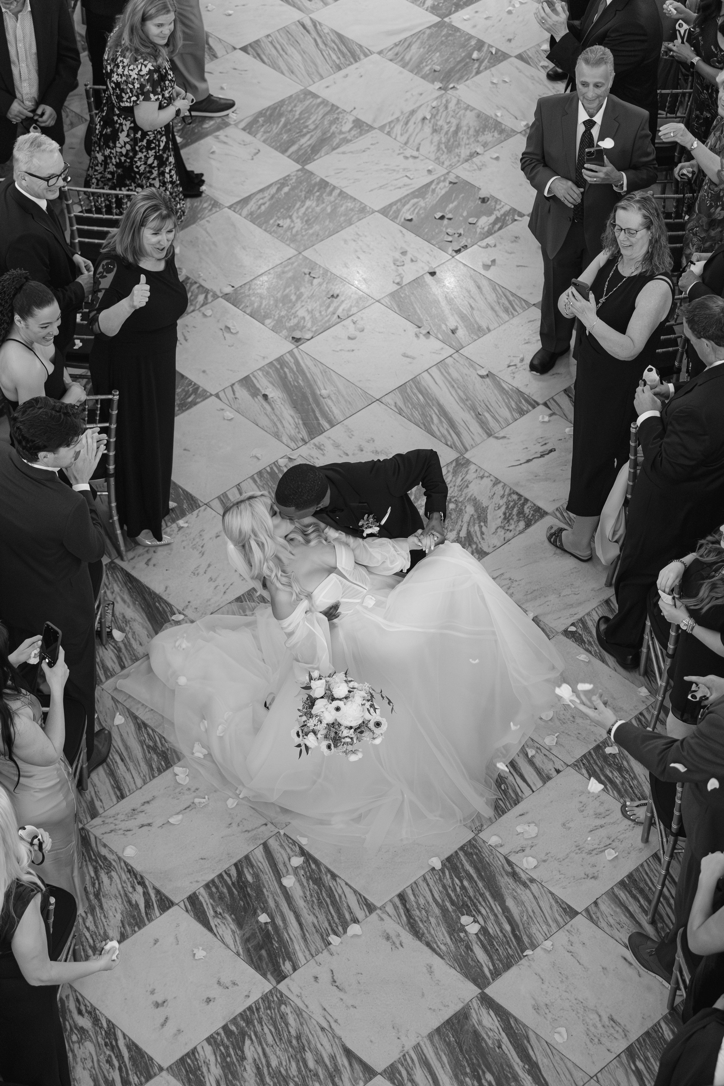 Overhead black and white view of a wedding couple on a checkered dance floor surrounded by guests at a reception.