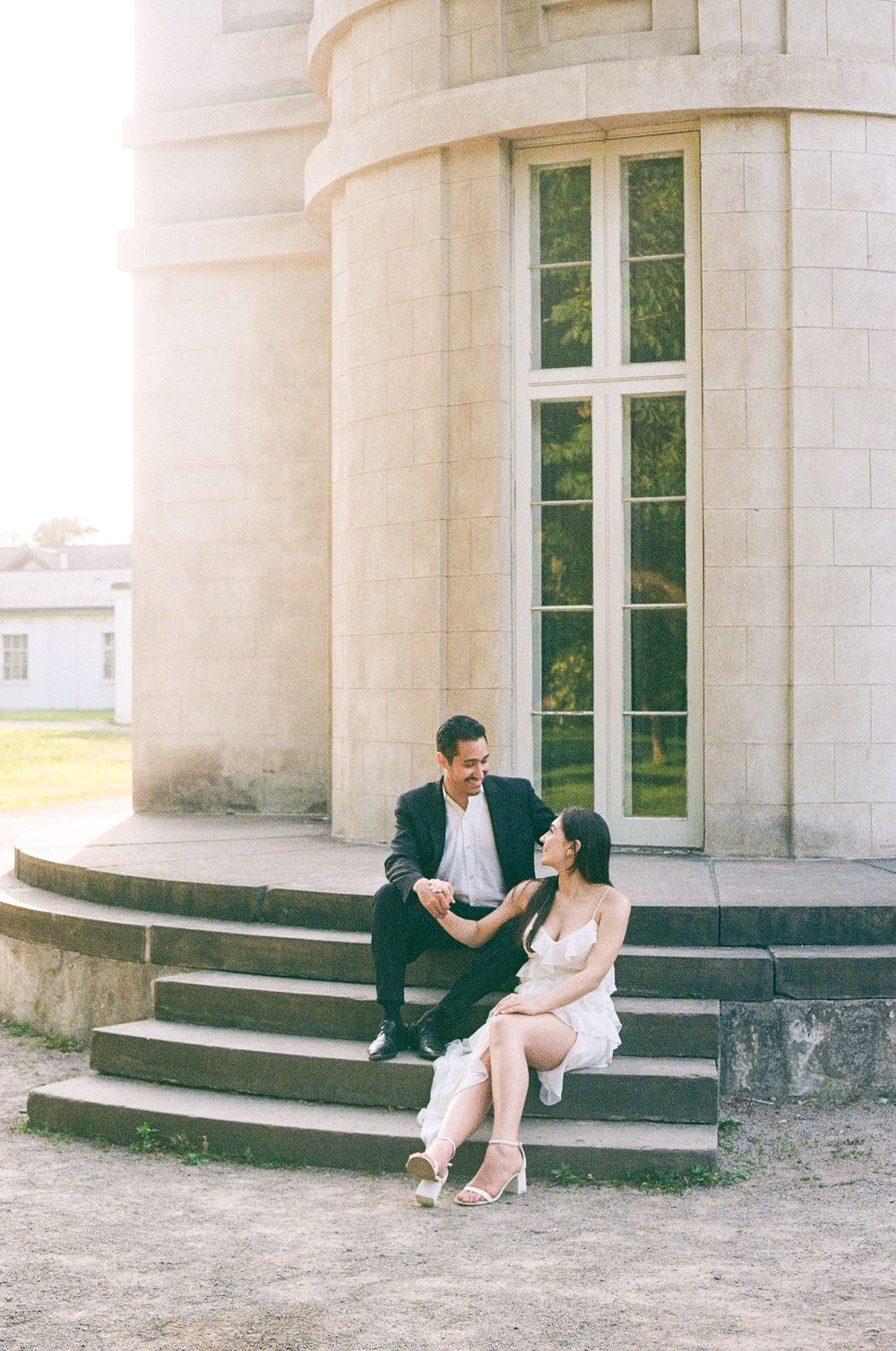 A couple sits intimately on stone steps beside a grand white building with tall windows.