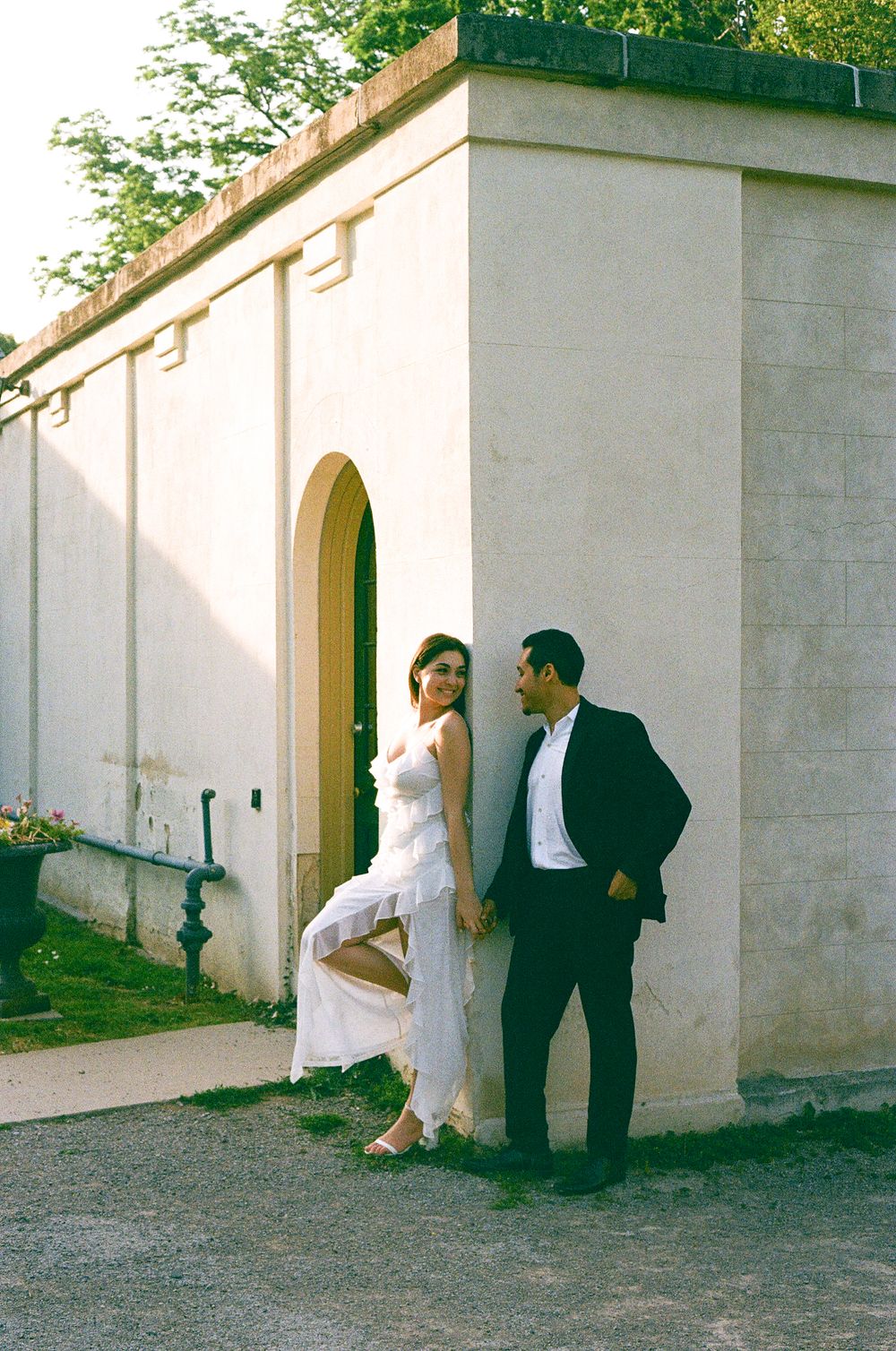 A couple in wedding attire stands by an arched doorway of a white stone building on a sunny day.