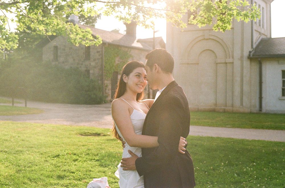 A couple embraces in formal wedding attire during golden hour with a stone building in the background.