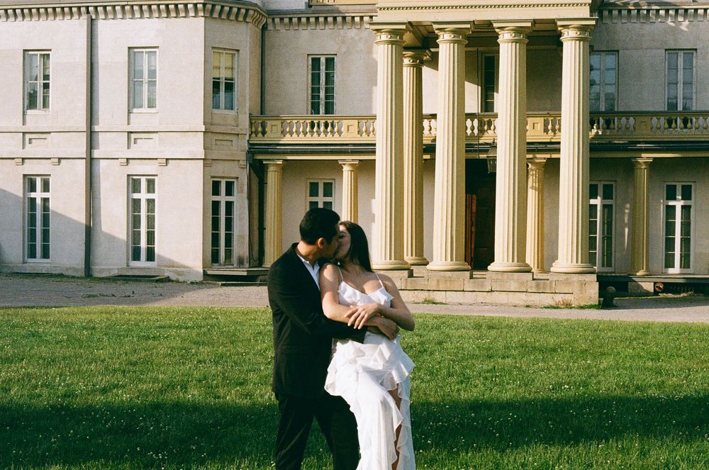 A couple in wedding attire embraces in front of an elegant white mansion with classical architecture and columns.
