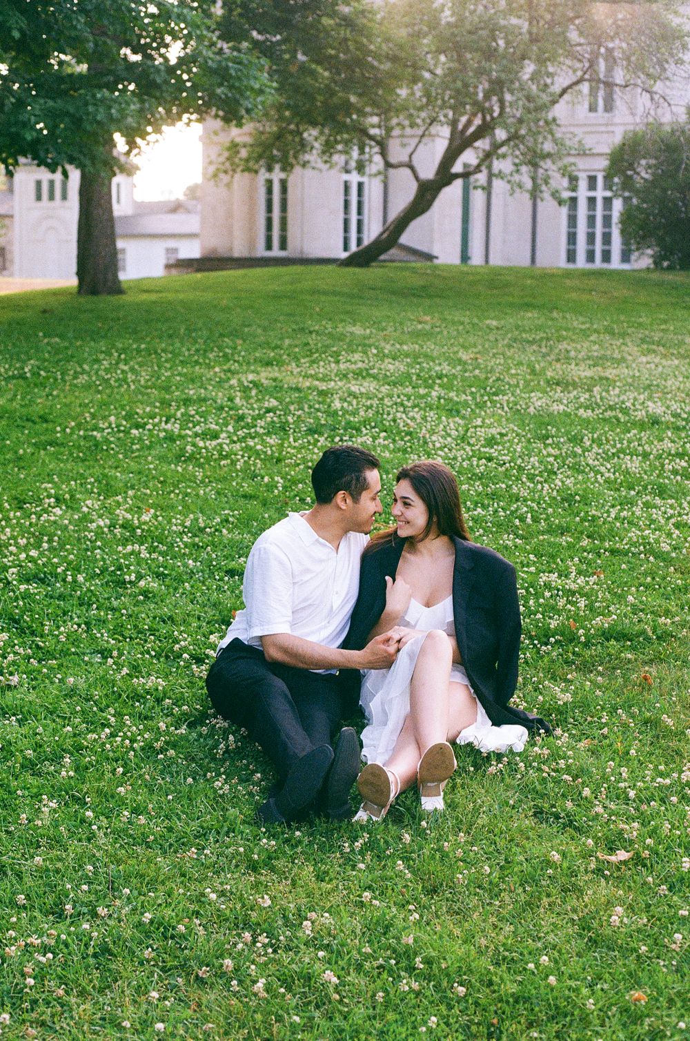 A romantic couple shares a tender moment while sitting on a grassy lawn near a stately building on a sunny day.