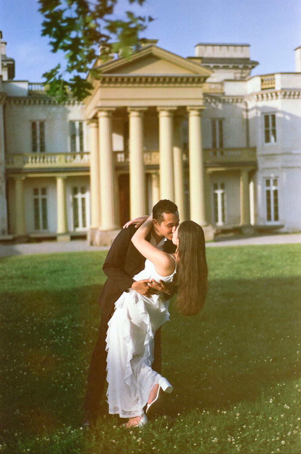 A romantic moment captured in front of a neoclassical mansion with white columns and manicured grounds.