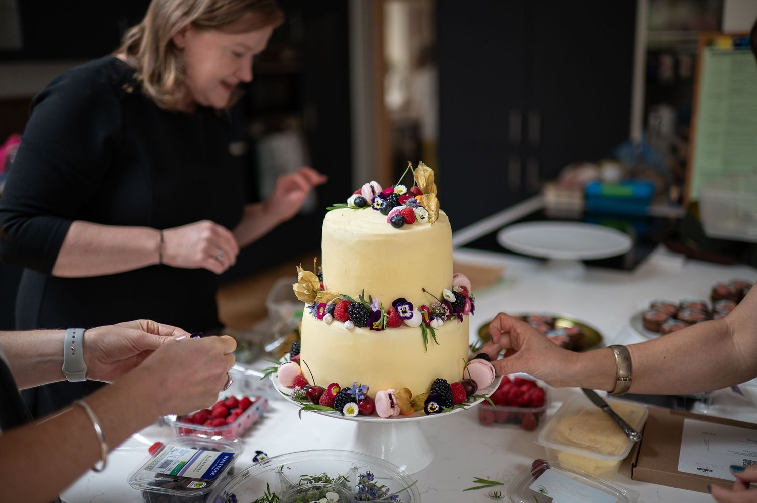 Two tier wedding cake with flowers and fruits, people are putting the finishing touches to it on a kitchen surface