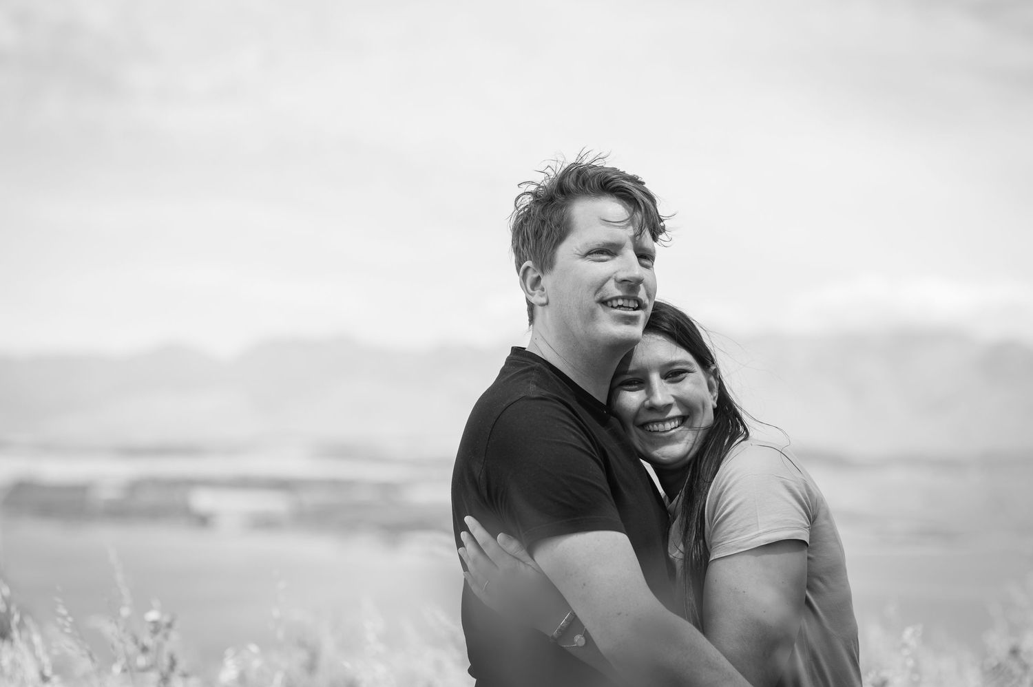 A romantic black and white portrait of a couple embracing outdoors against a blurred scenic background.