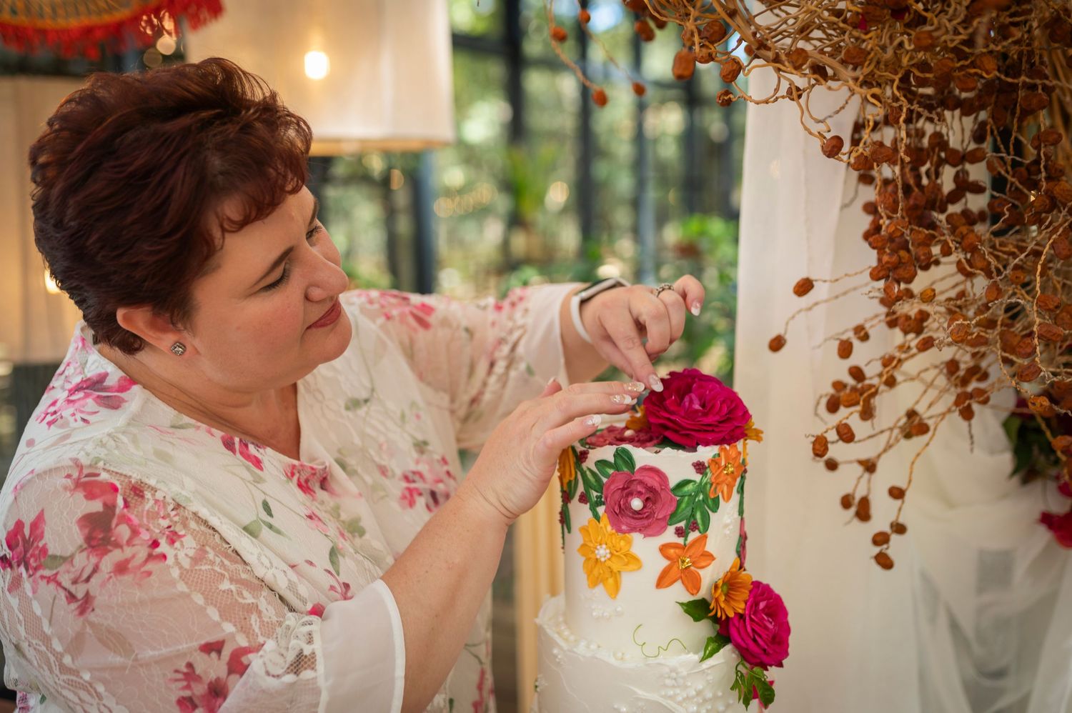 A baker wearing a floral print shirt delicately places vibrant flowers on a white wedding cake.