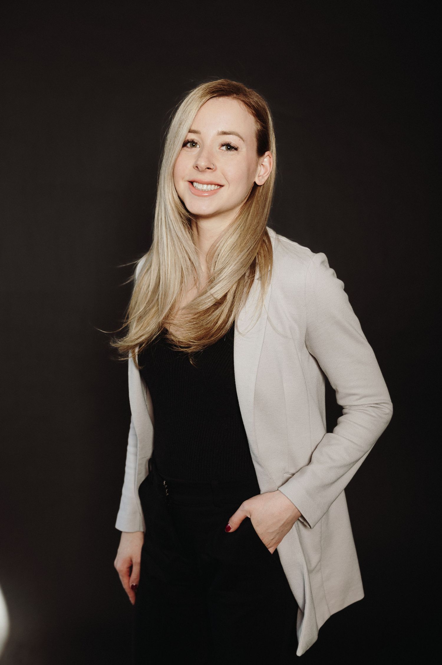 Professional studio portrait of a person wearing a light blazer over black top standing against dark background.