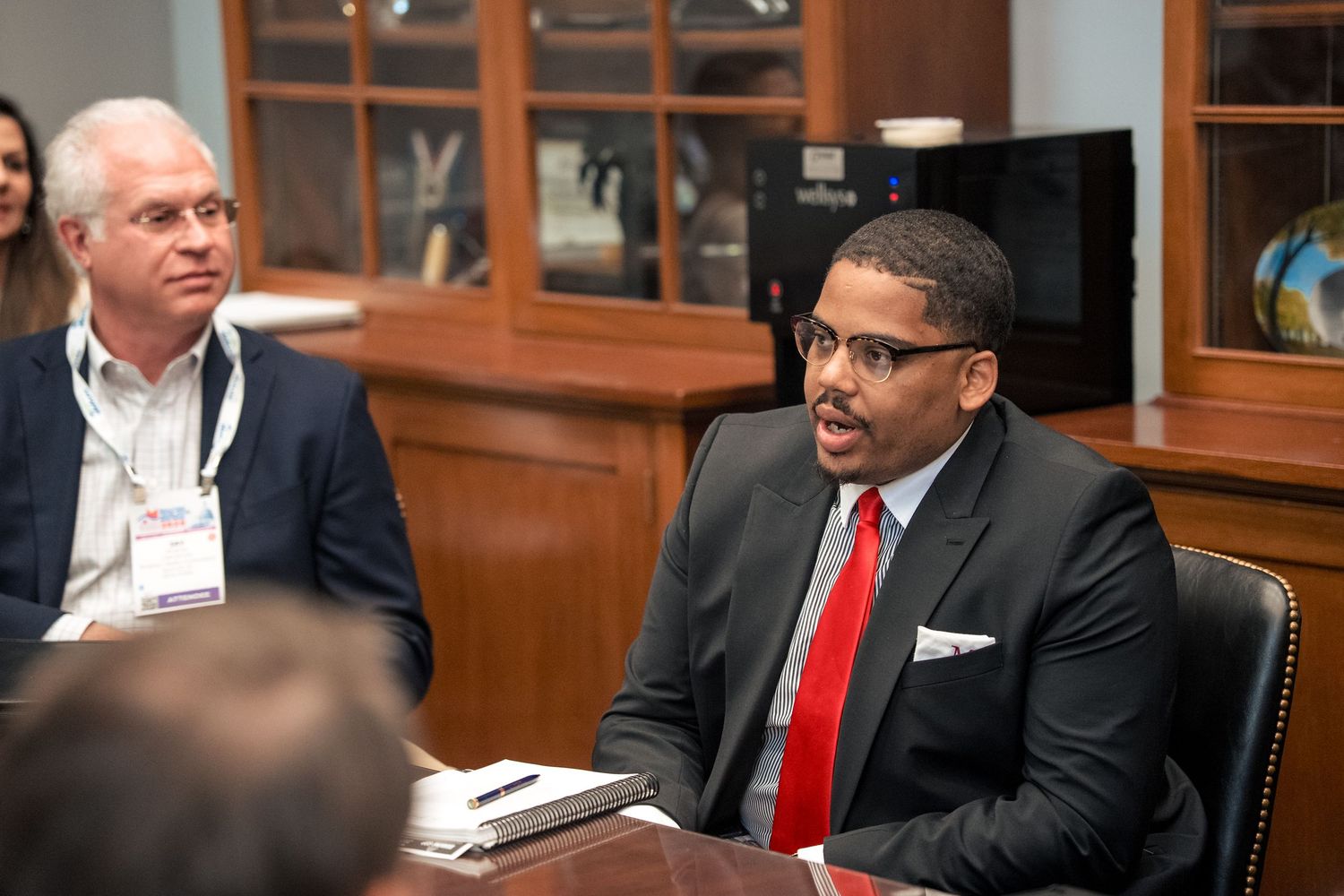 Four business professionals in suits sit at a conference table during a business meeting or discussion.