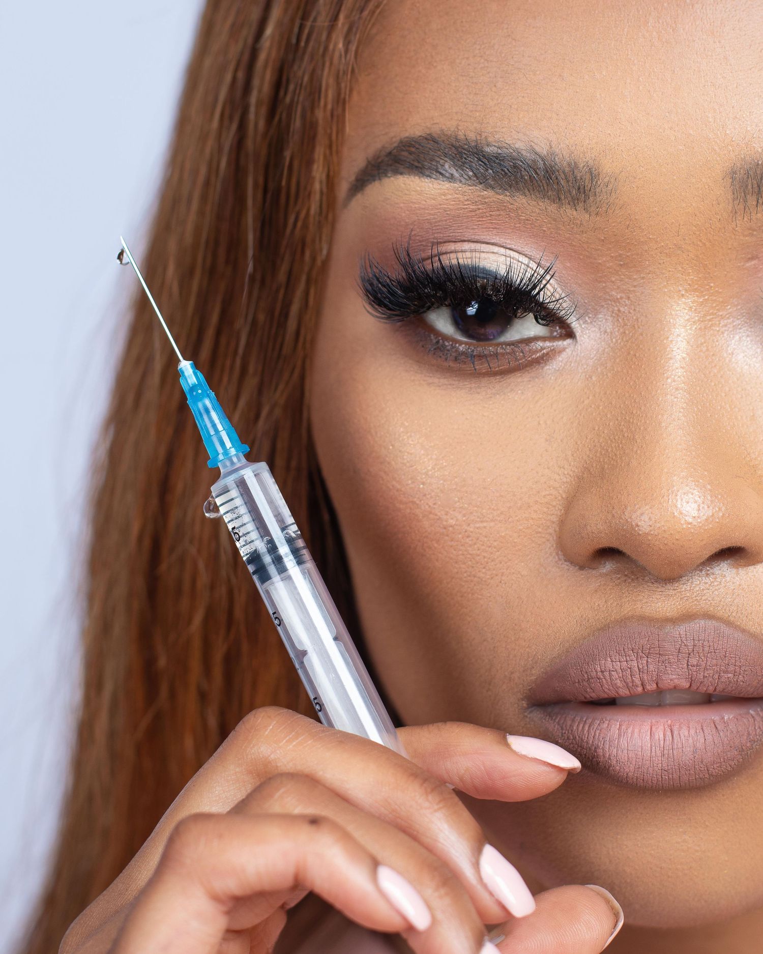 Close-up corporate beauty shot showing shimmery pink eyeshadow, winged liner and matte nude lips while holding a syringe.