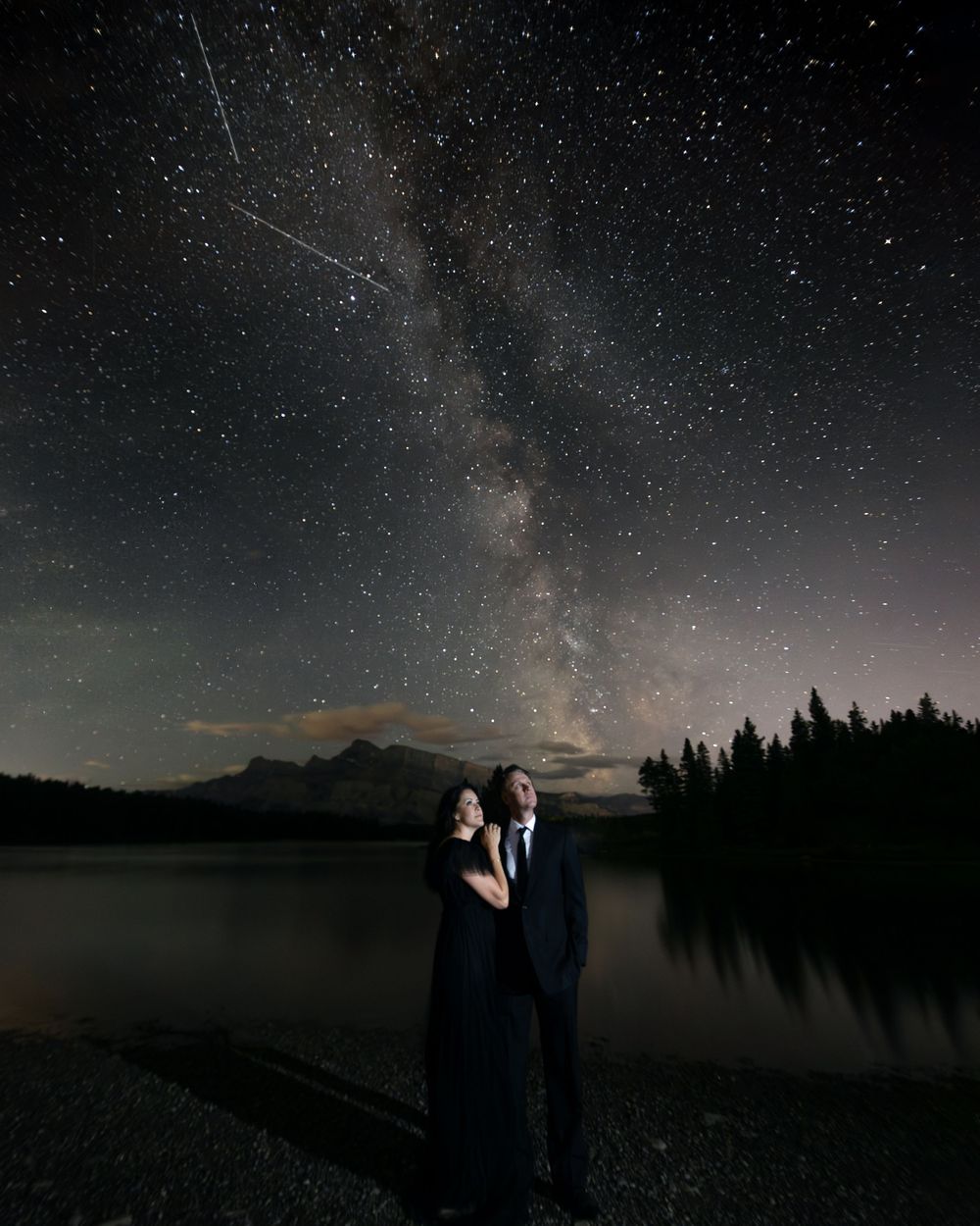 Silhouettes stand beneath the stunning Milky Way galaxy stretching across a starry night sky over a tranquil lake by Sofia Katherine.