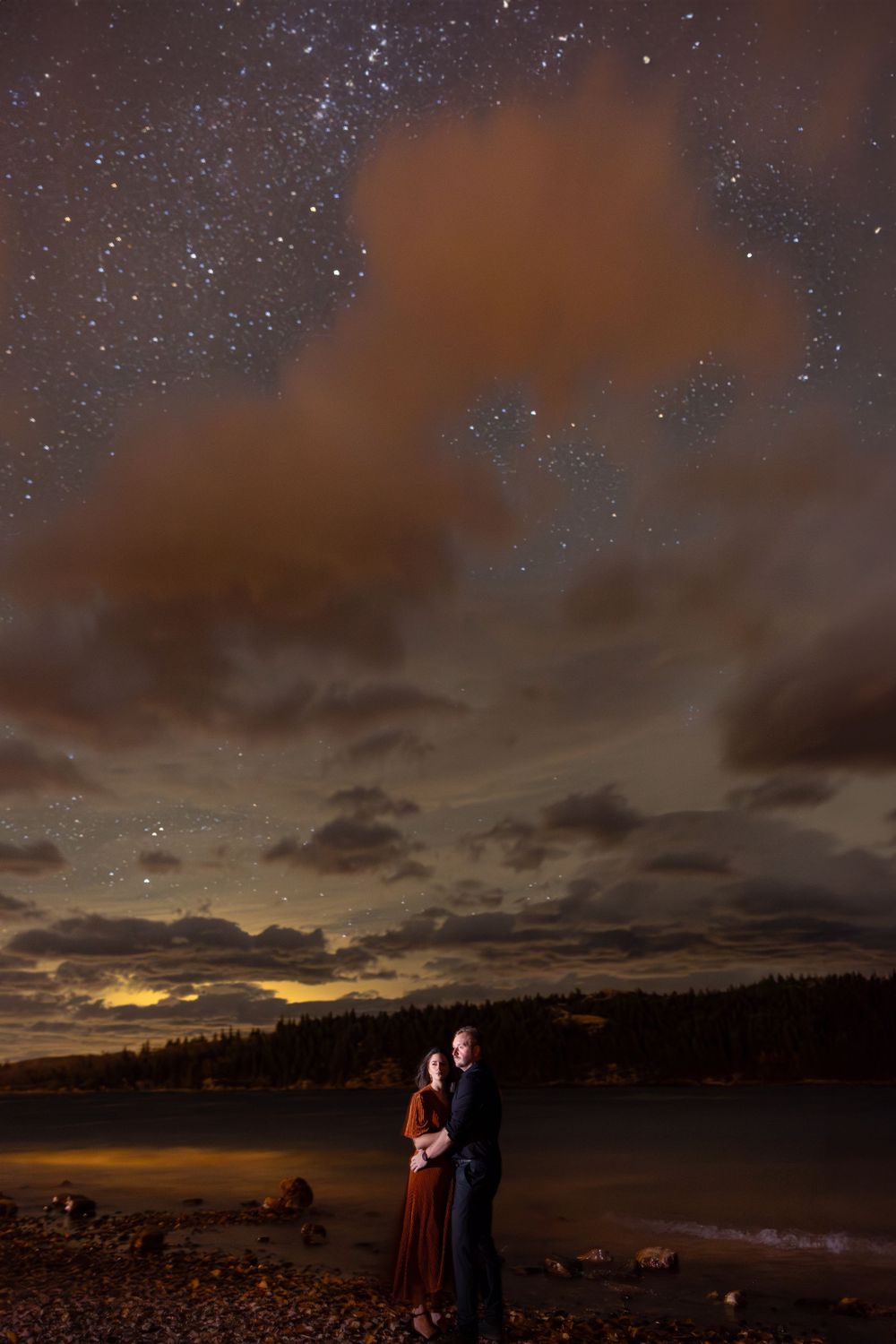 Couple stands together under a starry night sky with cloudy landscape and reflective lake in the background by Sofia Katherine Photography.