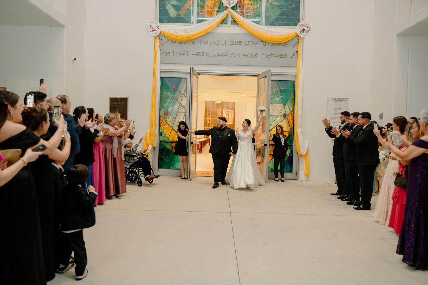 Wedding ceremony guests line both sides of a white hallway as the couple exits through ornately decorated doors.